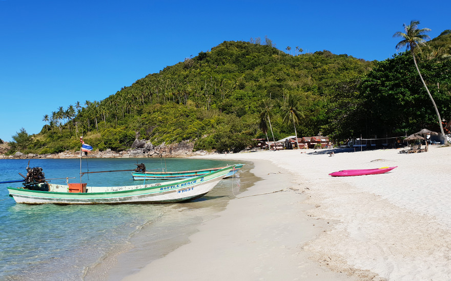 Bottle Beach im Norden von Koh Phangan mit weißem Sand, Longtailboot am Ufer, Palmen, bewaldeten Hügeln und türkisfarbenem Meer
