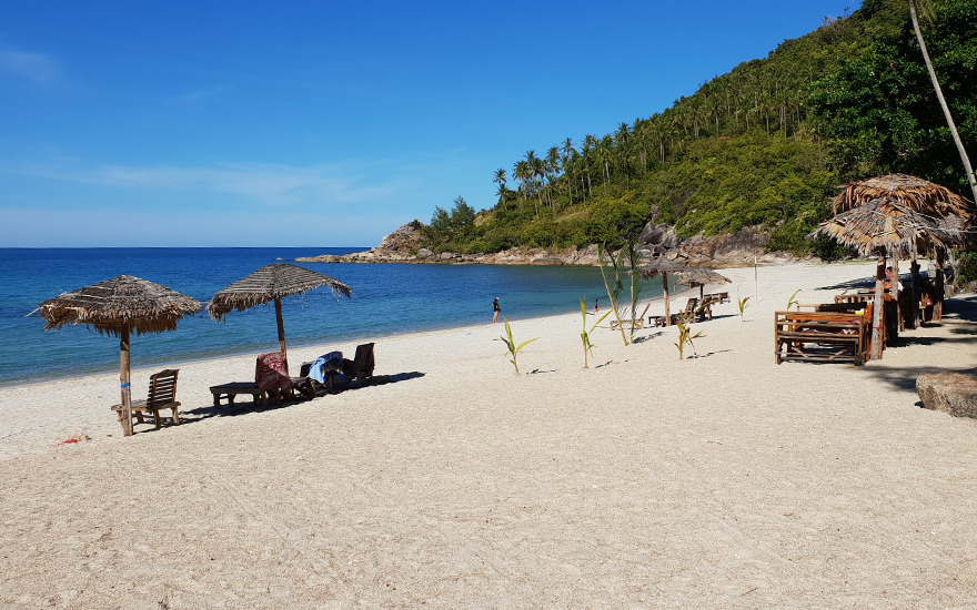 Bottle Beach mit einigen wenigen Sonnenschirmen und Liegen, weißem Sand und bewaldetem Hügel am Strandende