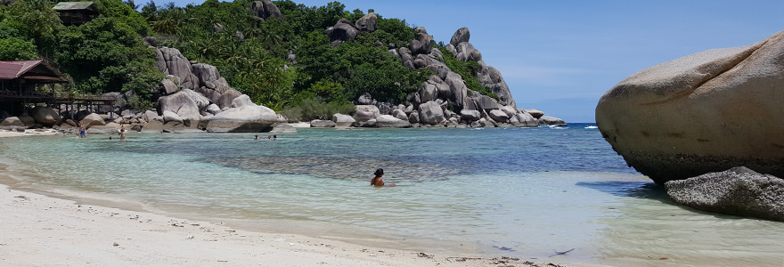 Freedom Beach mit seichtem, türkisfarbenem Meer, Badenden und bewaldeten Hügeln mit abgerundeten Felsen